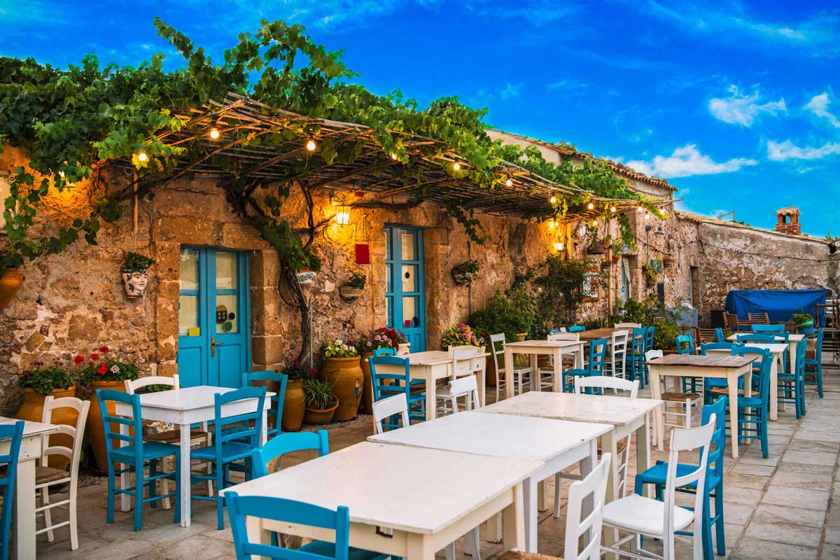 Outdoor dining area with tables and chairs under a stone building with blue doors and greenery.